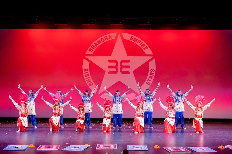 Kids and adults performing together on an indoor auditorium stage at the Bhangra Empire Dance Off