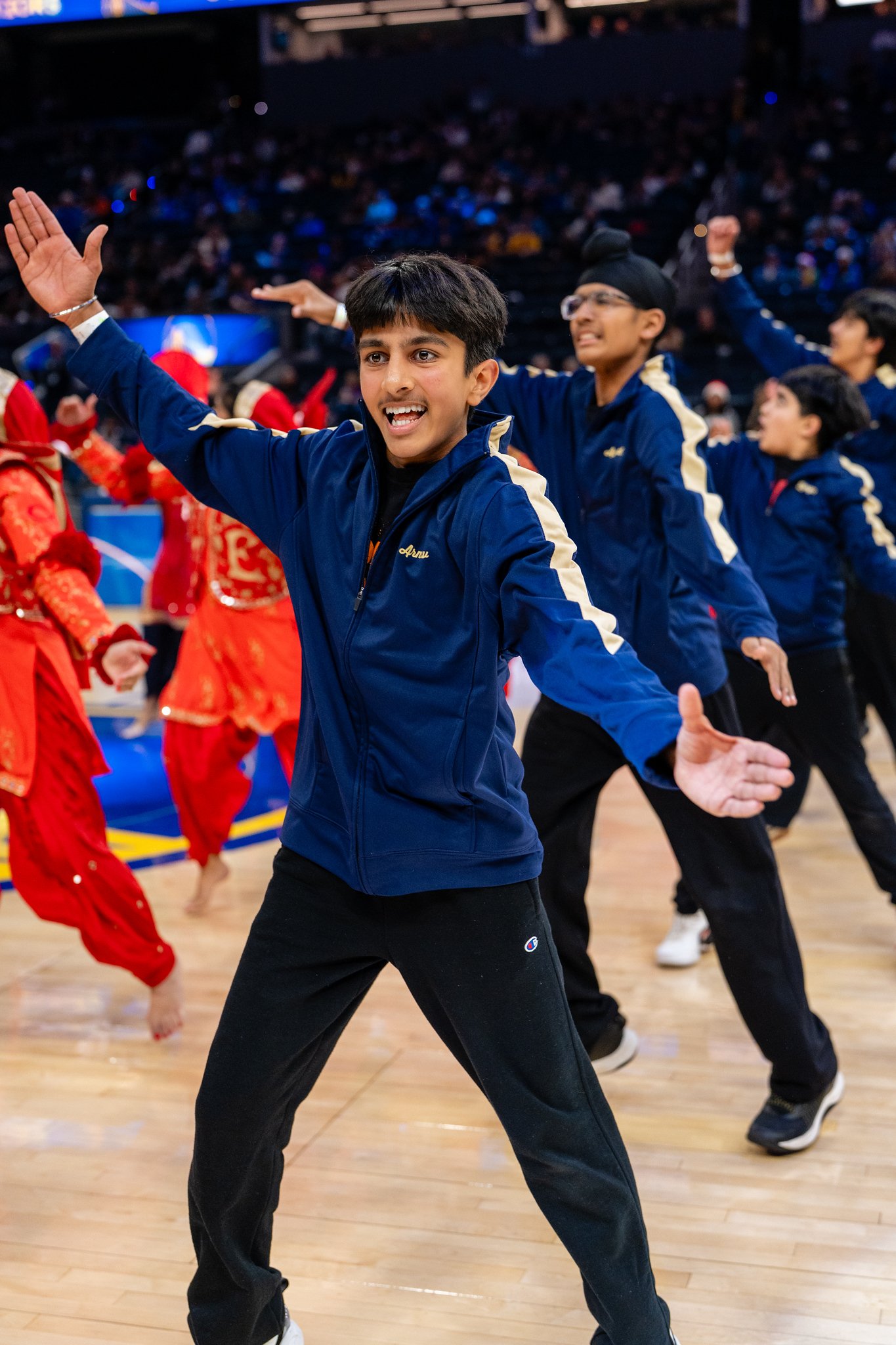 An Academy dancer in a blue jacket performing with arms outstretched on the Warriors court.