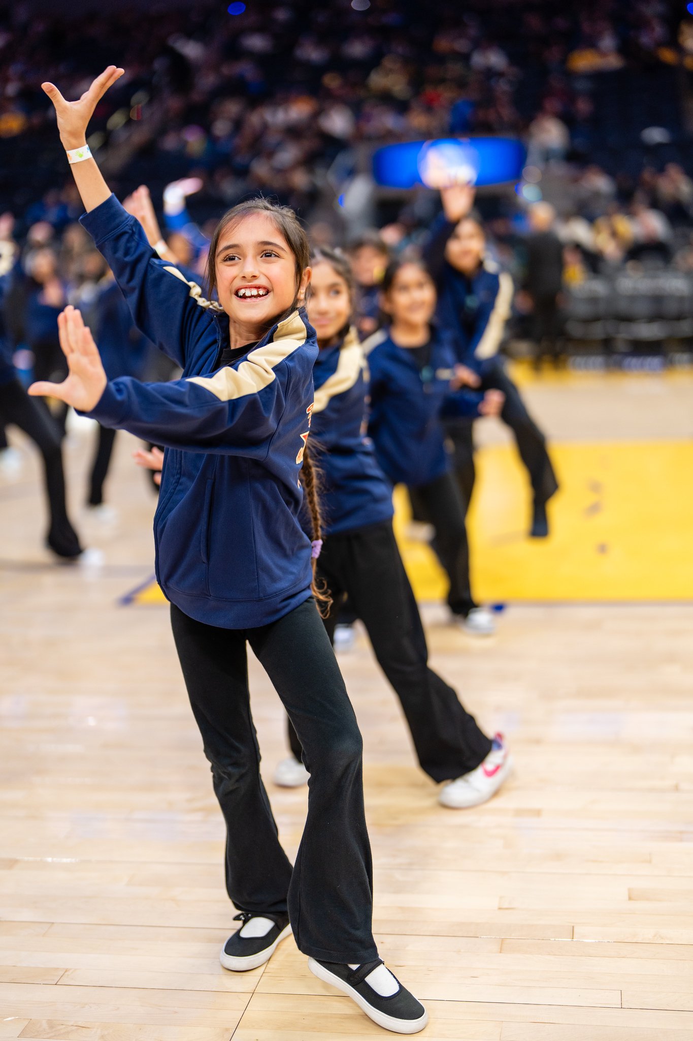 Young Academy dancers in blue jackets smiling and performing on the Warriors court.