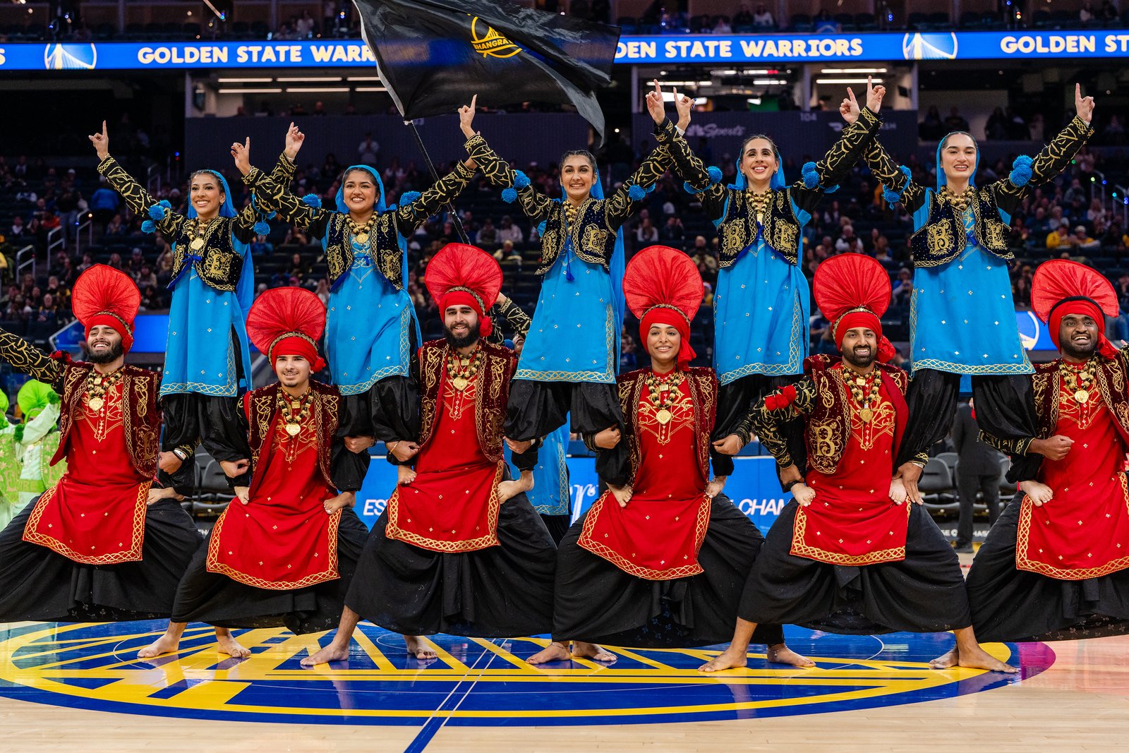 Bhangra Empire dancers in red and blue costumes celebrating on the Warriors court at Chase Center.